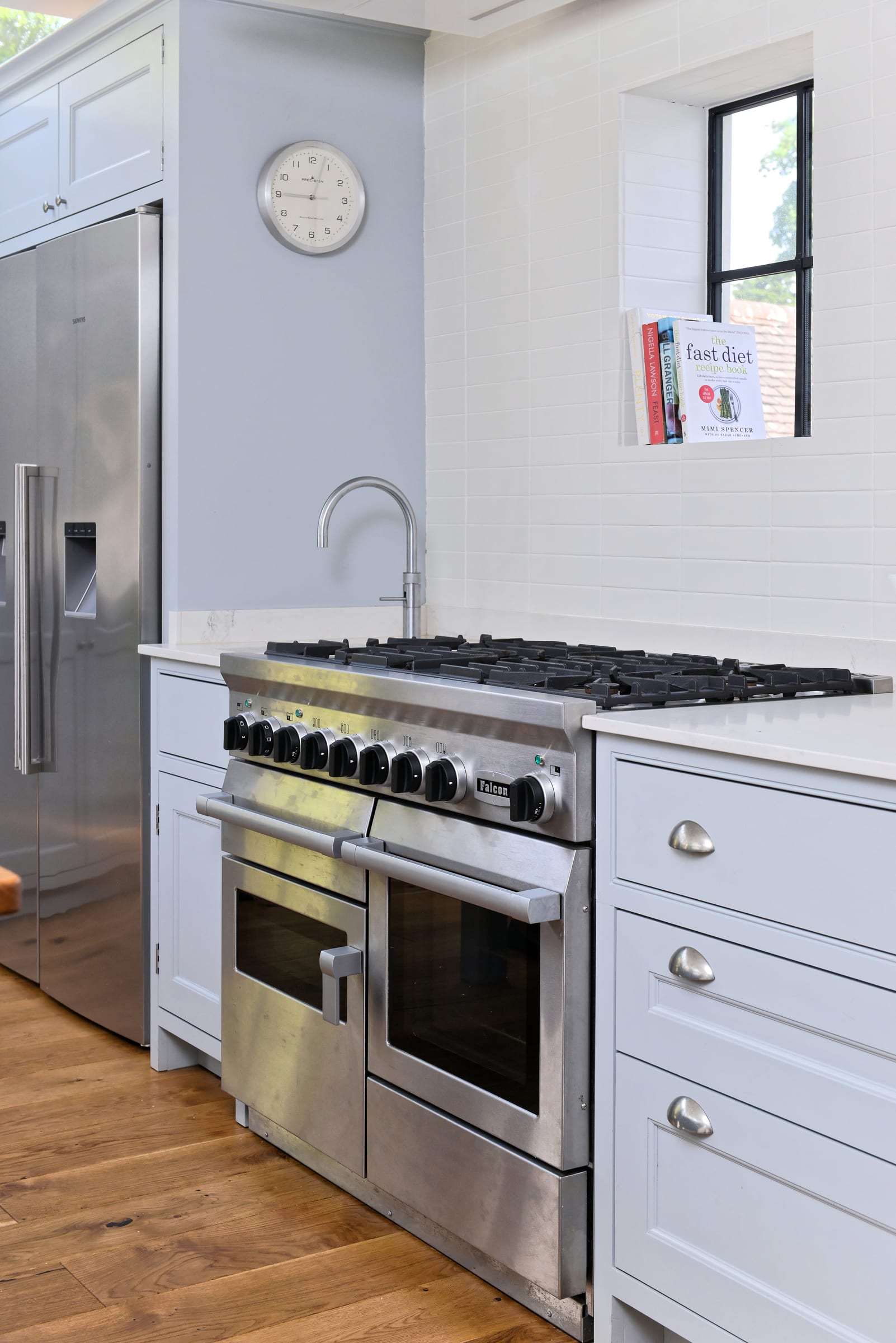 Pale blue in-frame cabinetry with a stainless steel range cooker and a cookbook nook by the window