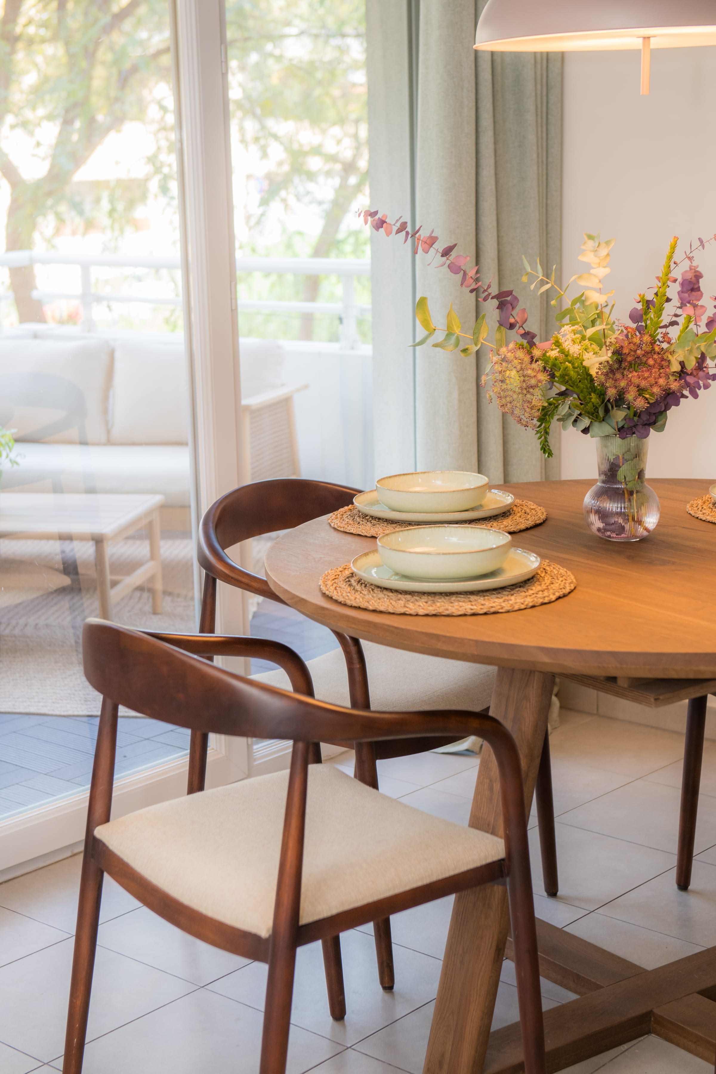 Dining table detail with wooden armchairs and a glass door opening onto a balcony with leafy views